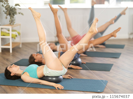 Together with students, teacher lies with fitball under her buttocks and performs swings with feet Together with students, teacher lies with fitball under her buttocks and performs swings with feet 115674037