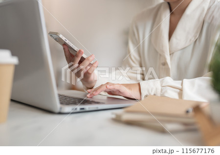 A businesswoman working remotely from a cafe, working on her laptop while using her smartphone. A businesswoman working remotely from a cafe, working on her laptop while using her smartphone. 115677176