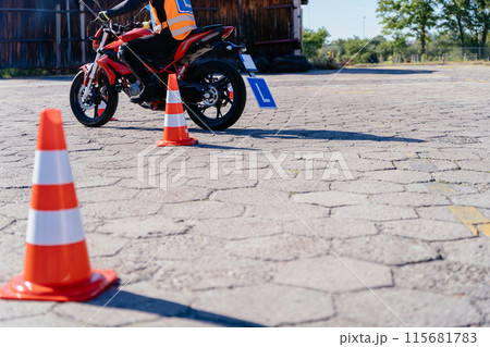 A woman on a motorcycle is riding past a row of orange cones. The cones are arranged in a line, and the man is approaching the middle of the row 115681783