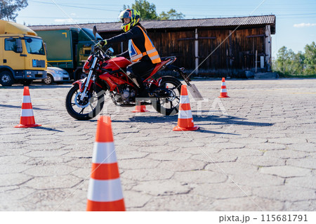 A man is riding a motorcycle in a parking lot with orange and white cones. The cones are arranged in a row, and the man is wearing a helmet A man is riding a motorcycle in a parking lot with orange and white cones. The cones are arranged in a row, and the man is wearing a helmet 115681791