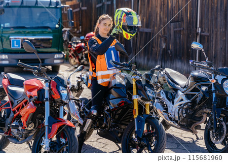 A woman puts a motorcycle helmet on motordrome with other motorcycles. Concept of motorcycle school of driving, as the woman is likely preparing for a ride or has just finished one. A woman puts a motorcycle helmet on motordrome with other motorcycles. Concept of motorcycle school of driving, as the woman is likely preparing for a ride or has just finished one. 115681960