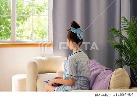 Teenage girl in blue pajamas looks out a window. 115684464