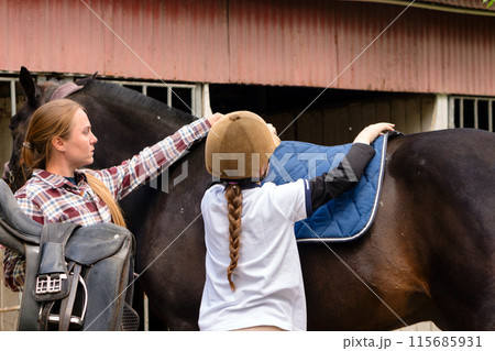 Instructor assisting girl to place saddle pad on horse 115685931