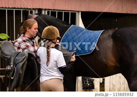 Instructor helping a girl position a saddle on a horse at a stable. 115685932