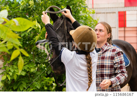 Woman and young rider collaboratively adjusting horse's bridle. Woman and young rider collaboratively adjusting horse's bridle. 115686167