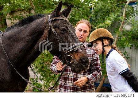 Woman and young rider during bridle adjustment on horse. Woman and young rider during bridle adjustment on horse. 115686171