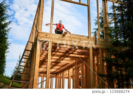 Carpenter constructing timber-framed two-story building. Man measures distances with measuring tape while dressed in workwear and hard hat. Ideology of contemporary, eco-friendly building practices. Carpenter constructing timber-framed two-story building. Man measures distances with measuring tape while dressed in workwear and hard hat. Ideology of contemporary, eco-friendly building practices. 115688274