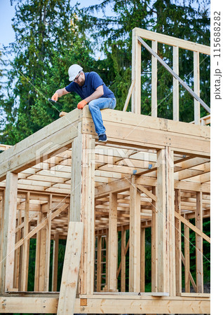 Carpenter building wooden, two-story frame house near woods. Bearded man with spectacles hammering nail while wearing protective helmet. The concept of contemporary ecological construction. Carpenter building wooden, two-story frame house near woods. Bearded man with spectacles hammering nail while wearing protective helmet. The concept of contemporary ecological construction. 115688282
