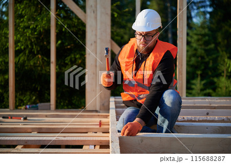 Wooden frame, two-story abode is being built by carpenter near forest. Bearded man wearing glasses is driving nails with a hammer, attired in a safety helmet and a construction vest. 115688287