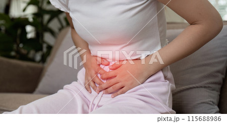 Woman Experiencing Stomach Pain While Sitting on a Couch in a Home Setting, Clutching Her Abdomen in Discomfort 115688986