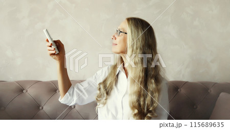 Mature woman turns on the air conditioner using the remote control sitting on couch in room at home 115689635