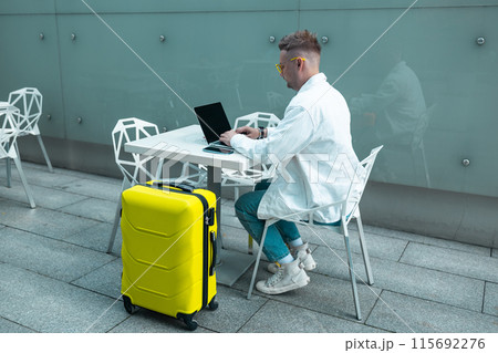 Happy traveling Caucasian man in stylish clothes uses mobile smartphone and laptop, checking trip destination on Internet. Male waiting at flight gates for plane boarding, smiling have a vacation trip 115692276