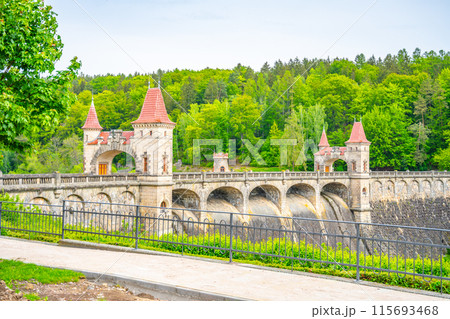 A view of the Les Kralovstvi Dam in Czechia, featuring the iconic castle-like structure with its arched bridge. The dam is surrounded by lush greenery and the water flows freely over the spillway. 115693468