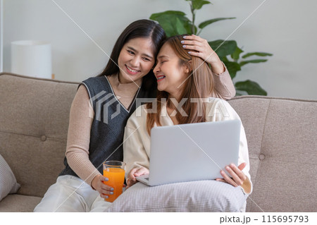 Happy Lesbian Couple Enjoying Healthy Orange Juice Together at Home While Relaxing on the Couch with a Laptop 115695793