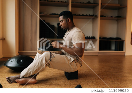 Side view of calm African-African man in trance playing meditative instrument tank drum using drumsticks sitting cross-legged on floor in meditation room. Concept of mindfulness, tranquility, harmony. 115699020