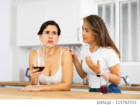 Upset woman drinking wine in kitchen while girlfriend consoling her 115702533
