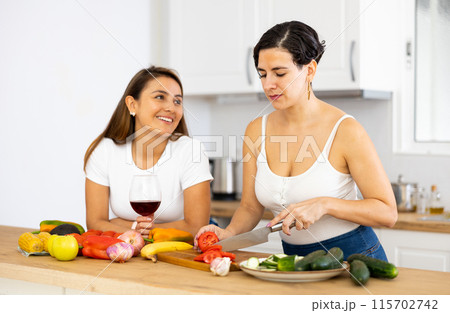 Two happy female friends preparing salad and drinking red wine together in kitchen Two happy female friends preparing salad and drinking red wine together in kitchen 115702742