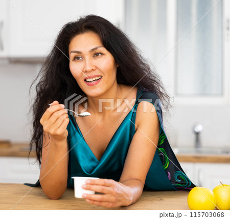Portrait of positive woman in nightdress eating yogurt at home 115703068
