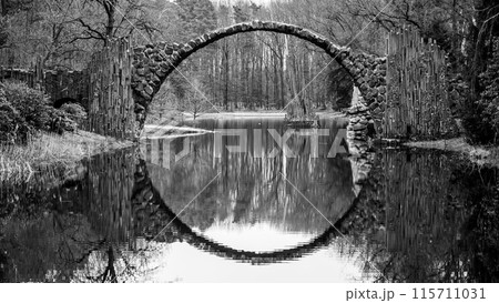 Rakotz Bridge, German: Rakotzbrucke, forms a perfect stone arch reflected in the calm waters of a serene German lake, surrounded by bare trees. Black and white image. 115711031