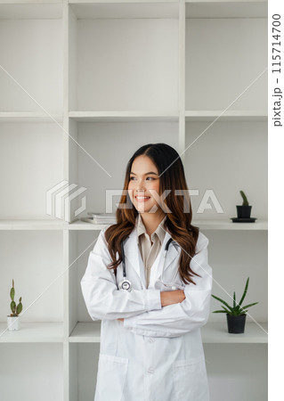 Professional female doctor in a white lab coat standing confidently with arms crossed in a modern office setting with shelves and plants. Professional female doctor in a white lab coat standing confidently with arms crossed in a modern office setting with shelves and plants. 115714700