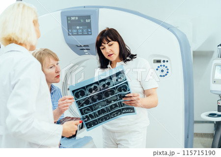 A woman is looking at a medical image of a woman's head. Two females doctor wearing a white lab coat and is standing next to a woman in a blue shirt A woman is looking at a medical image of a woman's head. Two females doctor wearing a white lab coat and is standing next to a woman in a blue shirt 115715190