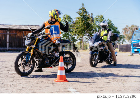 Motorcycle driving school concept. Charming woman in orange vest with Letter sign is riding a motorcycle in a parking lot. Instructor man on the left is wearing a yellow vest. 115715290