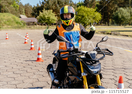 Positive female student wearing a helmet, taking motorcycle lessons and practicing riding, points her finger at the letter L on her vest. Student exam in motorcycle school. Positive female student wearing a helmet, taking motorcycle lessons and practicing riding, points her finger at the letter L on her vest. Student exam in motorcycle school. 115715527