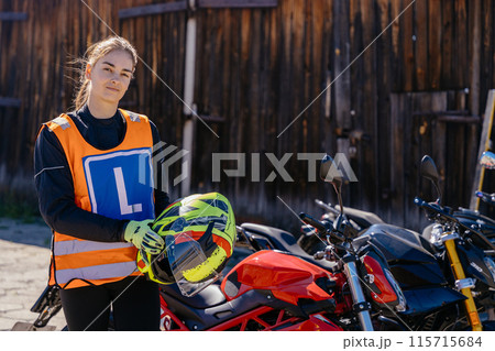 Moto school track driving school. Portrait of female student dressed in an orange vest displaying the letter L holds helmet before motorcycle lessons and practicing ride at moto school. Moto school track driving school. Portrait of female student dressed in an orange vest displaying the letter L holds helmet before motorcycle lessons and practicing ride at moto school. 115715684