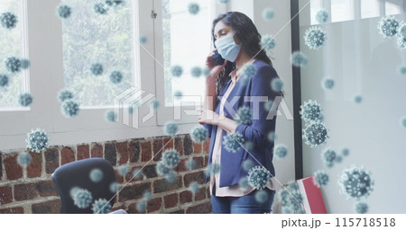 Image of covid 19 cells floating over man wearing face mask, using smartphone in office 115718518
