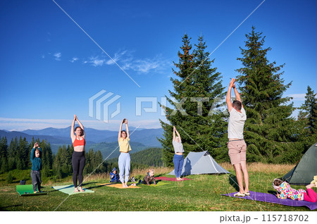 Group of people doing yoga pose outdoor in camping in the mountains. Adults and children standing on yoga mats, each doing yoga pose under clear blue sky in morning. 115718702