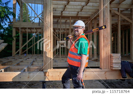 Carpenter building wooden frame two-story house. Bearded man in glasses holding a level, wearing protective helmet, overalls and orange vest. Concept of modern ecological construction. Carpenter building wooden frame two-story house. Bearded man in glasses holding a level, wearing protective helmet, overalls and orange vest. Concept of modern ecological construction. 115718764