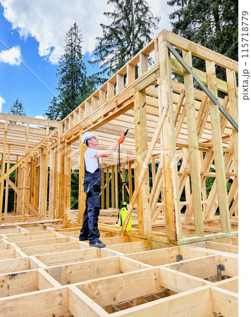 Laborer constructing wooden frame house near forest. Man treating woods, applying fire retardant using sprayer, while dressed in protective suit, helmet. Concept of modern eco-friendly construction. Laborer constructing wooden frame house near forest. Man treating woods, applying fire retardant using sprayer, while dressed in protective suit, helmet. Concept of modern eco-friendly construction. 115718779