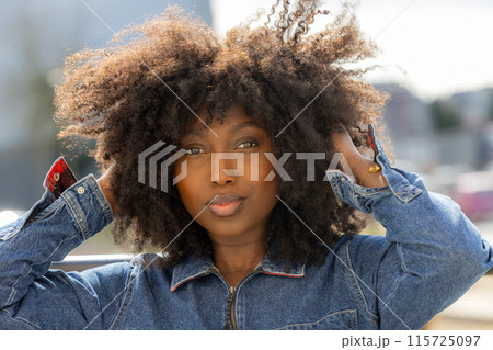 A charming woman with afro hair and a denim jacket appears attractive while outdoors A charming woman with afro hair and a denim jacket appears attractive while outdoors 115725097