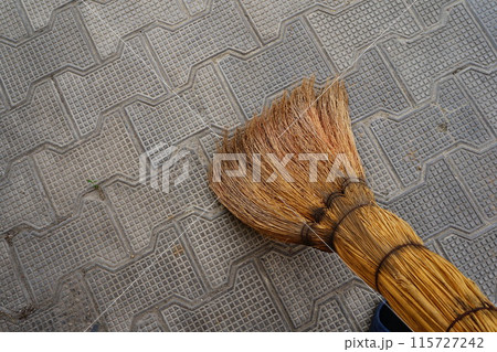 A brown natural broom made of plants sweeps the textured pavement paved with cobblestones. Maintaining the cleanliness of the house space, entrances and courtyard A brown natural broom made of plants sweeps the textured pavement paved with cobblestones. Maintaining the cleanliness of the house space, entrances and courtyard 115727242