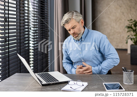 Businessman experiencing stomach pain while working on a laptop at a modern office desk. Stress, health issues, and discomfort. 115728371