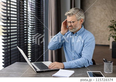 Middle-aged man with glasses and beard experiencing head pain while working on laptop at office desk. Concept of work stress and headache. 115728374