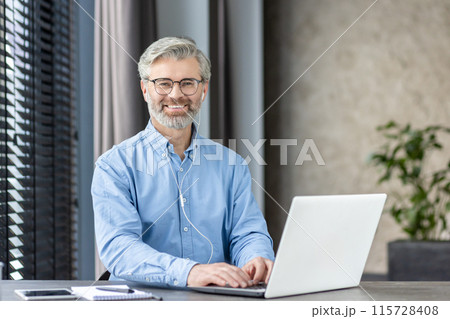 Smiling mature man with glasses working on laptop in modern office environment, exuding confidence and professionalism. 115728408