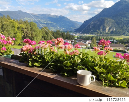 Idyllic view from balcony with geranium flowers and coffee cup in the village of Wiesing towards green valley with Alps and Lake Achensee in Tyrol, Austria. Summer sunny day. 115728423