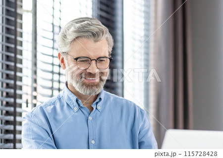 Smiling mature businessman wearing glasses and blue shirt, working on laptop with white blinds in the background. Smiling mature businessman wearing glasses and blue shirt, working on laptop with white blinds in the background. 115728428