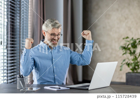 Happy businessman celebrating career success at work in office environment with a laptop and documents. 115728439