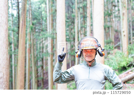 仕事場の山林をバックに右手人差し指を上に指差し微笑んでいるフォレストワーカー 115729050