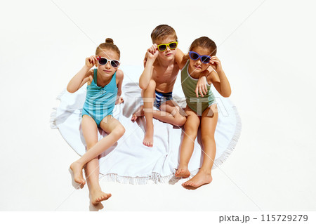 Cute little children, girls and boy in swimsuits relaxing on beach towel against white studio background. 115729279