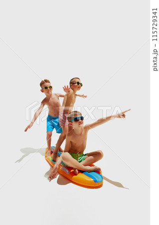 Three boys, dressed in vibrant swim trunks and cool sunglasses, stand on inflatable surfboard, pretending to ride waves against white studio background. 115729341