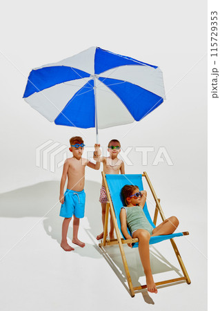 Three children enjoy a summer day. One boy holds a blue-and-white beach umbrella, another boy stands beside him, and girl relaxes in beach chair Three children enjoy a summer day. One boy holds a blue-and-white beach umbrella, another boy stands beside him, and girl relaxes in beach chair 115729353