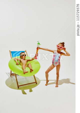 Kids have fun on sunny day. Boy relaxes in beach chair with green inflatable ring, while his friend, girl holds toy water blaster. Kids have fun on sunny day. Boy relaxes in beach chair with green inflatable ring, while his friend, girl holds toy water blaster. 115729376
