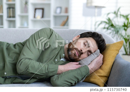 Close-up photo of a young tired man sleeping and resting at home on the couch, lying with his eyes closed, holding a pillow in his hands. 115730740