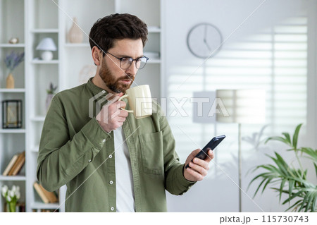 A young man in glasses and a green shirt is standing at home, holding a cup of drink and using a mobile phone. 115730743