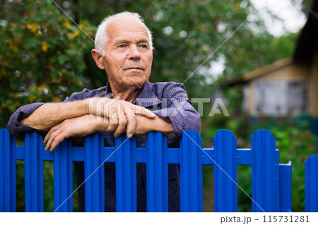 Portrait of gray-haired owner at fence of his country house in village Portrait of gray-haired owner at fence of his country house in village 115731281
