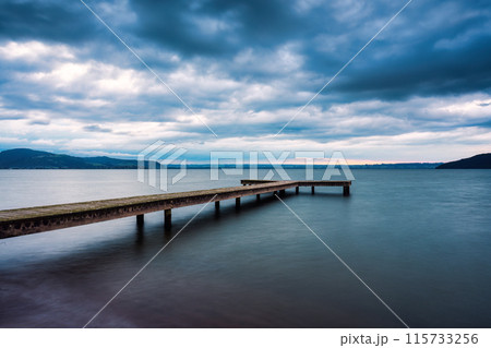 Wooden pier leading into the lake with cloudy sky in gloomy day Wooden pier leading into the lake with cloudy sky in gloomy day 115733256