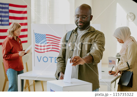 Confident young Black male citizen placing ballot paper into ballot box at polling place on presidential election day 115735322